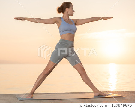 Active experienced woman teacher performs yoga asana on mat on seashore at sunset. Concept of healthy lifestyle 117949670