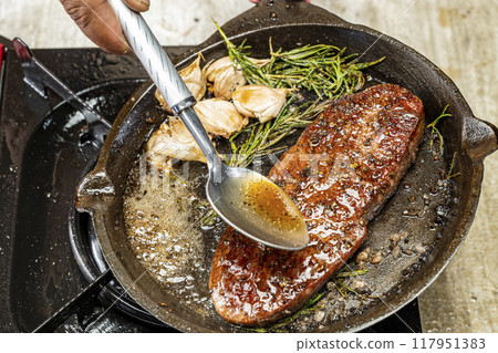 A person is cooking a sirloin steak in a pan with herbs and spices A person is cooking a sirloin steak in a pan with herbs and spices 117951383