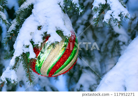 Large Christmas ball of geometric pattern with yellow, red, green stripes is hanging on the spruce tree branch covered with snow. 117952271