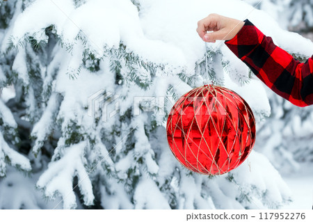 A hand in red plaid shirt is holding a large red Christmas ball to decorate outside, snowy branches on the background. A hand in red plaid shirt is holding a large red Christmas ball to decorate outside, snowy branches on the background. 117952276
