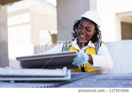 An African American female contractor is examining rooftop tiles at the construction site. The female foreman is inspecting the construction materials. An African American female contractor is examining rooftop tiles at the construction site. The female foreman is inspecting the construction materials. 117953806