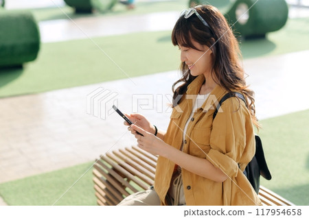 traveler, young Asian woman in her 30s, smiles brightly at a vibrant Thai station, smartphone in hand. Capture the joy of connected solo exploration with this captivating portrait. 117954658