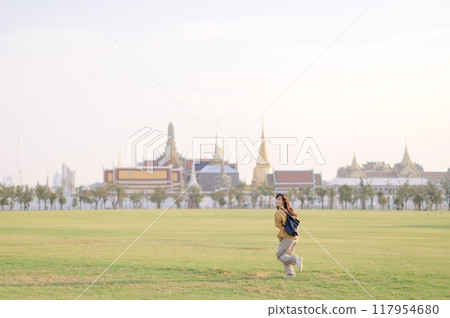 Traveler asian woman in her 30s, backpack slung over her shoulder, explores Wat Pra Kaew with childlike wonder. Sunlight dances on the golden rooftops, and a carefree smile Traveler asian woman in her 30s, backpack slung over her shoulder, explores Wat Pra Kaew with childlike wonder. Sunlight dances on the golden rooftops, and a carefree smile 117954680