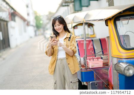 Young Asian woman backpack traveler standing a side of Tuk Tuk taxi on summer vacations at Bangkok, Thailand. Journey trip lifestyle, Asia summer tourism concept. Young Asian woman backpack traveler standing a side of Tuk Tuk taxi on summer vacations at Bangkok, Thailand. Journey trip lifestyle, Asia summer tourism concept. 117954712