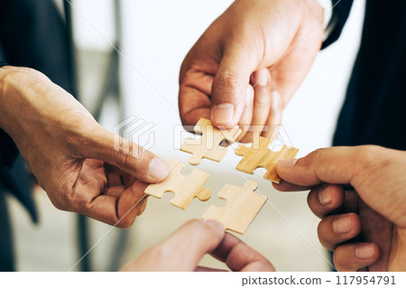 The concept of teamwork and partnership. Businessman's hands putting together puzzle pieces in the office and join together in a jigsaw puzzle team. Charity. Volunteers. Business unity as a team. The concept of teamwork and partnership. Businessman's hands putting together puzzle pieces in the office and join together in a jigsaw puzzle team. Charity. Volunteers. Business unity as a team. 117954791
