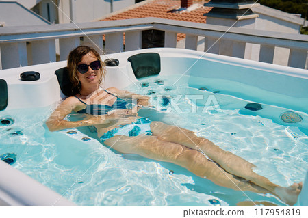 Woman wearing sunglasses and swimsuit relaxing in outdoor hot tub with bubbling water and enjoying sunny day 117954819