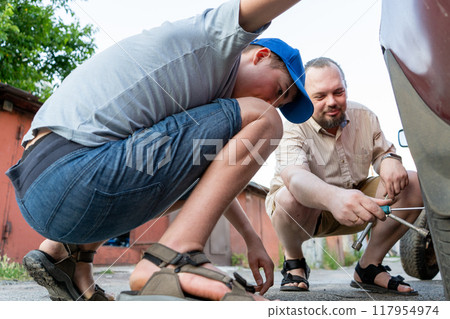 Dad and teenage son work together on the street near the garage. Outdoor activity. Family spend time. Helping and teach. Replacing an old wheel and tire on a car Dad and teenage son work together on the street near the garage. Outdoor activity. Family spend time. Helping and teach. Replacing an old wheel and tire on a car 117954974
