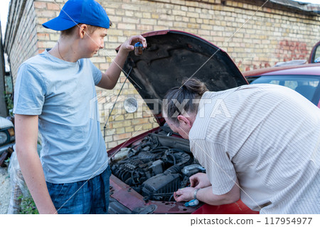 Dad and teenage son work together on the street near the garage. Outdoor activity. Family spend time. Helping and teach. The car hood is open. Engine repair and oil change. Dad and teenage son work together on the street near the garage. Outdoor activity. Family spend time. Helping and teach. The car hood is open. Engine repair and oil change. 117954977