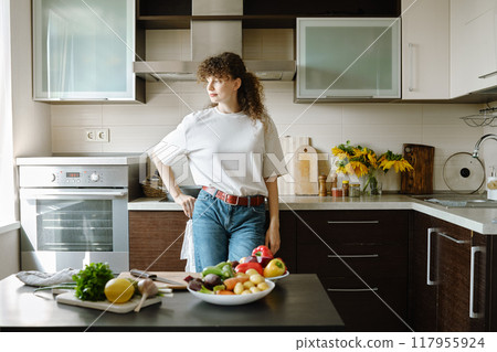 Woman stands in a kitchen with fresh vegetables on the table and looking to the window Woman stands in a kitchen with fresh vegetables on the table and looking to the window 117955924