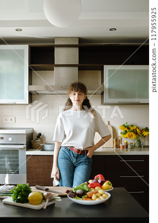 Woman stands in a brightly lit kitchen with a fresh herbs and vegetables on the table Woman stands in a brightly lit kitchen with a fresh herbs and vegetables on the table 117955925