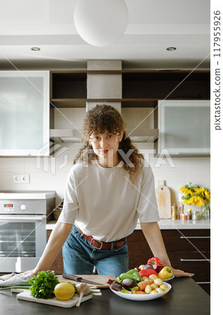 Curly-haired woman leaning against a kitchen table with organic vegetables on it Curly-haired woman leaning against a kitchen table with organic vegetables on it 117955926
