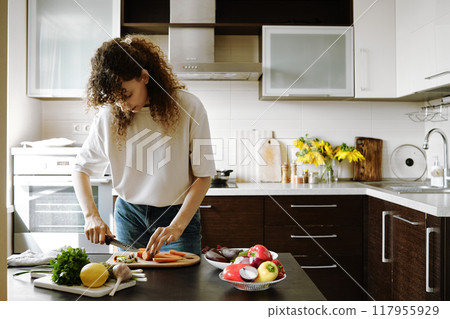 Curly-haired woman cutting on half fresh carrot in the kitchen Curly-haired woman cutting on half fresh carrot in the kitchen 117955929