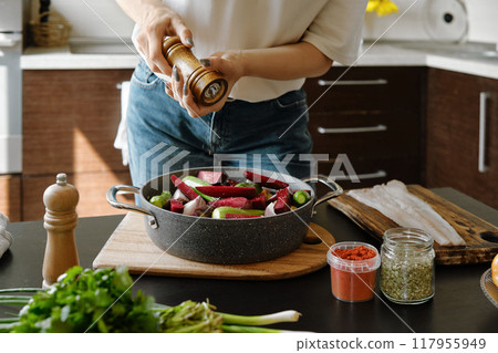 Close-up view of hands sprinkle pepper from manual spice mill on vegetables in a baking pan Close-up view of hands sprinkle pepper from manual spice mill on vegetables in a baking pan 117955949