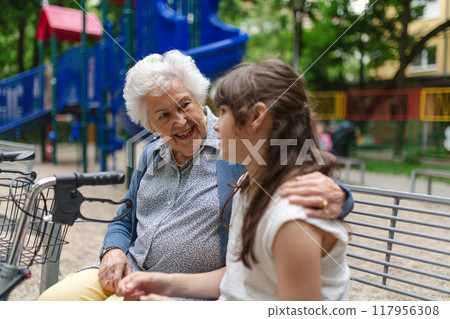 Grandma with walker spending time with granddaughter in city park, sitting on bench at children playground, talking. After school. 117956308