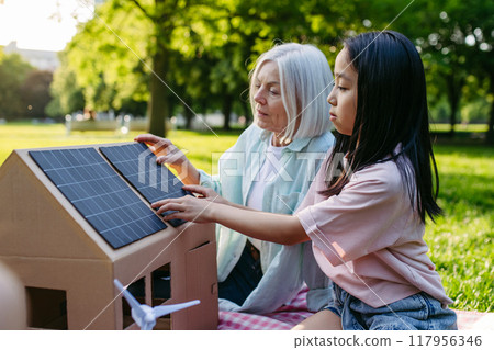 Girl learning about renewable energy and solar panels during sustainable education class outdoors, using cardboard model of house wit solar panel on roof. 117956346