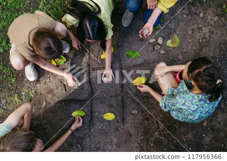 Students playing tic-tac-toe with stones and leaves, grid in soil. Outdoor sustainable education class. 117956366