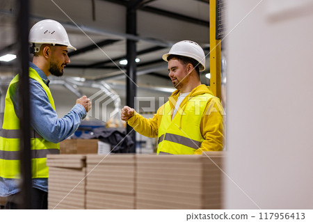 Young man with Down syndrome working in modern factory, fist bump with colleague. Concept of workers with disabilities, support in workplace. Young man with Down syndrome working in modern factory, fist bump with colleague. Concept of workers with disabilities, support in workplace. 117956413