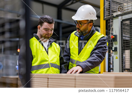 Young man with Down syndrome working in modern factory, colleague teaching him about his job, showing process. Concept of workers with disabilities, support in workplace. Young man with Down syndrome working in modern factory, colleague teaching him about his job, showing process. Concept of workers with disabilities, support in workplace. 117956417