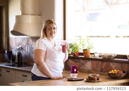Overweight woman in the kitchen, making fresh smoothie. Choosing healthy food, drinking smoothie from glass. 117956523
