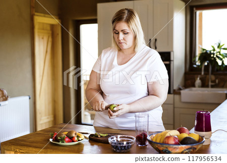 Overweight woman in the kitchen, making fresh smoothie. Choosing healthy food, cutting fruit on cutting board. 117956534