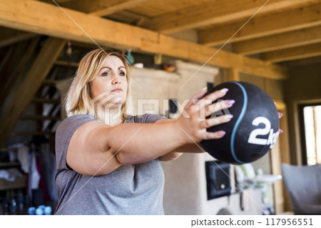 Overweight woman exercising at home, using weighted medicine ball. Workout in living room. 117956551