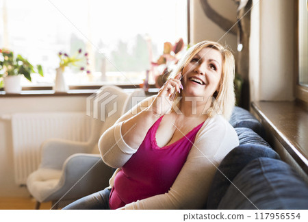 Portrait of beautiful young overweight woman at home sitting on sofa making phone call on smartphone. 117956554