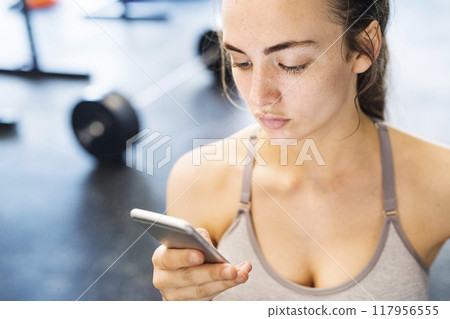 Young fitness girl checking her performance on smatphone after hard workout in gym. Young fitness girl checking her performance on smatphone after hard workout in gym. 117956555