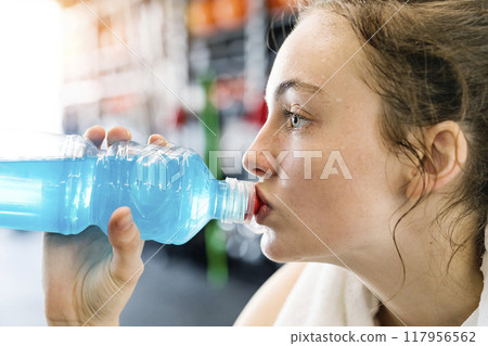 Young fitness girl drinking blue sport drink after hard workout in gym, side view. Young fitness girl drinking blue sport drink after hard workout in gym, side view. 117956562