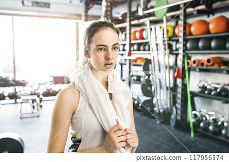 Young fitness girl with dental braces feeling exhausted after hard workout in gym, wiping her face with towel. 117956574