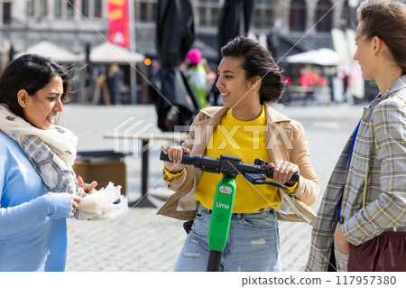 A Group of Three Friends Enjoying a Fun Scooter Ride and Street Food in the Bustling City Square 117957380