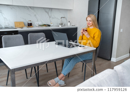 A woman is shopping online from her modern kitchen using a laptop and smartphone A woman is shopping online from her modern kitchen using a laptop and smartphone 117959126