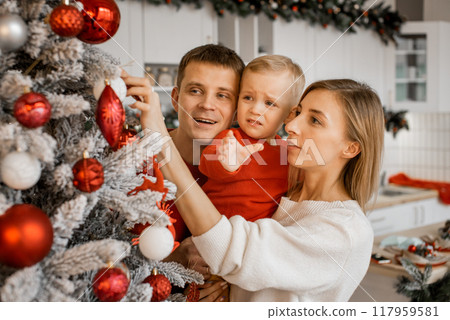 Happy mother, father and little son decorating christmas tree together at home, Little boy is crying and wants to take toy ball in his arm Happy mother, father and little son decorating christmas tree together at home, Little boy is crying and wants to take toy ball in his arm 117959581