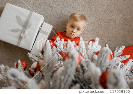 Top view of a cute little boy in red sweater lying on the floor under the Christmas tree next to wrapped Chrismas presents 117959590