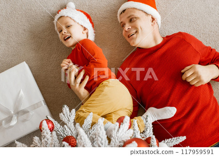 Top view of happy joyful family dad and son in santa hats playing and having fun on the floor under christmas tree. Christmas celebration at home. 117959591