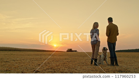 A farmer couple chats in a field at sunset, with their dog beside them and a combine harvester working in the distance. 117960253