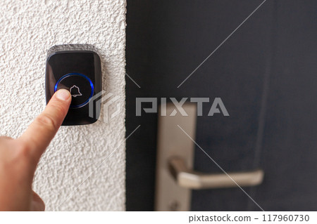 Pressing Black Doorbell with Blue Ring on White Wall 117960730