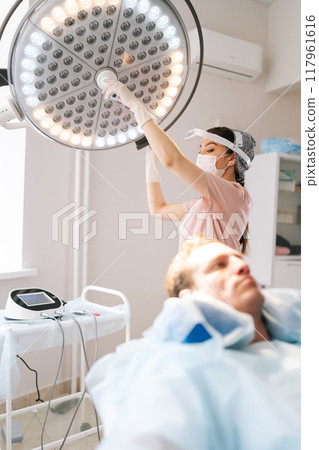 Vertical selective focus shot of female nurse in medical uniform adjusting light preparation performing medical procedure at clinic. Surgeon adjusting surgical lamp before medical operation. 117961616