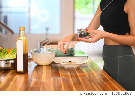 Shot of young woman in activewear carefully adding dried fruits from a small jar into a bowl, preparing a healthy desserts Shot of young woman in activewear carefully adding dried fruits from a small jar into a bowl, preparing a healthy desserts 117961968
