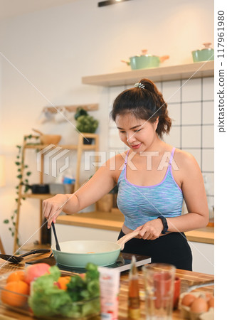 Young woman stirring ingredients in a pan on the stove, preparing a healthy meal in a modern kitchen 117961980