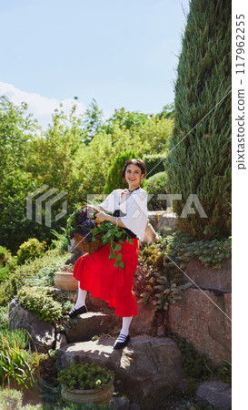 Woman in traditional outfit, holding basket of grapes and lemons, stands gracefully on rocky path surrounded by lush greenery, exuding rustic charm. Concept of vintage, art. Woman in traditional outfit, holding basket of grapes and lemons, stands gracefully on rocky path surrounded by lush greenery, exuding rustic charm. Concept of vintage, art. 117962255