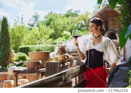 Smiling in sunlight, woman holds up glass of red wine, her outfit and surroundings evoking charm on idyllic countryside vineyard. Concept of vintage, art. 117962256