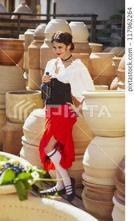 Woman with playful smile on her face, clutches bottle of wine, rich colors of dress contrasting with muted pottery tones. Concept of vintage, art. Woman with playful smile on her face, clutches bottle of wine, rich colors of dress contrasting with muted pottery tones. Concept of vintage, art. 117962264