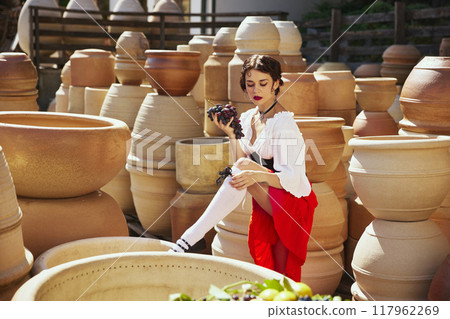 Grapes in hand, lady in thoughtful pose, surrounded by intricate designs of pottery, embodying essence of harvest. Concept of vintage, art. Grapes in hand, lady in thoughtful pose, surrounded by intricate designs of pottery, embodying essence of harvest. Concept of vintage, art. 117962269