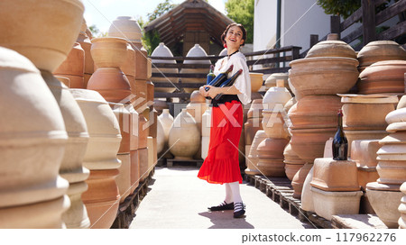 Standing amidst large clay pots, woman balances several wine bottles in her arms, her face glowing with bright, contented smile. Concept of tourism and art. 117962276