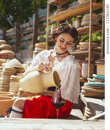 Elegant woman, dressed traditional outfit, seated amidst large clay pots, she gracefully pours wine from earthenware jug into glass. Concept of tourism and art. 117962280