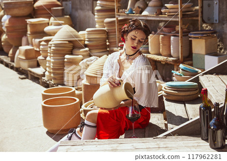 Spanish woman, dressed traditional outfit, elegant girl, Seated amidst large clay pots, she gracefully pours wine from earthenware jug into glass. Concept of tourism and art. 117962281