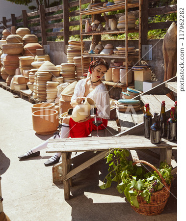 Seated amidst large clay pots, woman dressed historical clothes gracefully pours wine from earthenware jug into glass. Concept of tourism and art. Seated amidst large clay pots, woman dressed historical clothes gracefully pours wine from earthenware jug into glass. Concept of tourism and art. 117962282