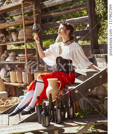 Beautiful, elegant woman dressed in historical attire sits on wooden bench, holding glass of wine up to sunlight. Concept of autumn and harvesting. 117962304