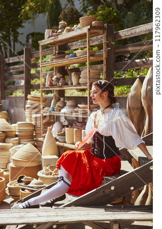 Beautiful young woman with dark hair, dressed in red skirt and white blouse, relaxes with fan in rustic pottery workshop. Concept of autumn and harvesting. Beautiful young woman with dark hair, dressed in red skirt and white blouse, relaxes with fan in rustic pottery workshop. Concept of autumn and harvesting. 117962309