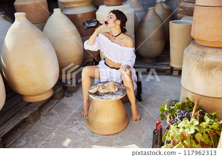 Beautiful young woman working clay in pottery studio, and drinks wine, surrounded by earthy-toned jars. Concept of autumn and harvesting. Beautiful young woman working clay in pottery studio, and drinks wine, surrounded by earthy-toned jars. Concept of autumn and harvesting. 117962313